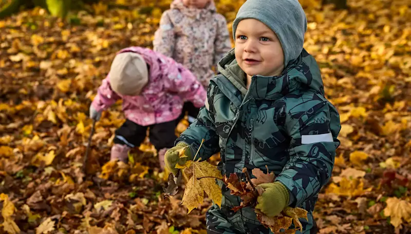 Barn i fargerike vinterdresser leker med høstløv. Barnet i forgrunnen har grønne votter og svarte vintersko synlige blant gule løv.