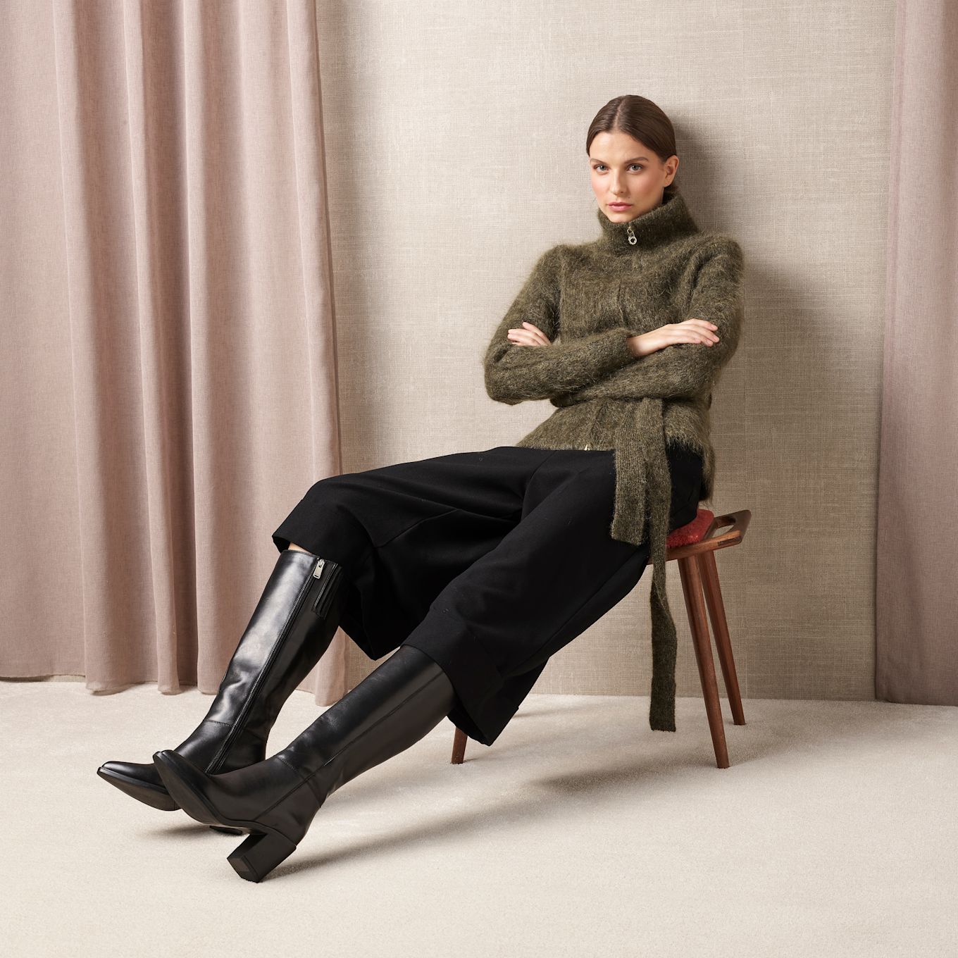 Model i grøn sweater og sorte bukser med elegante sorte læderstøvler med hæl og lynlås på siden, siddende på taburet.