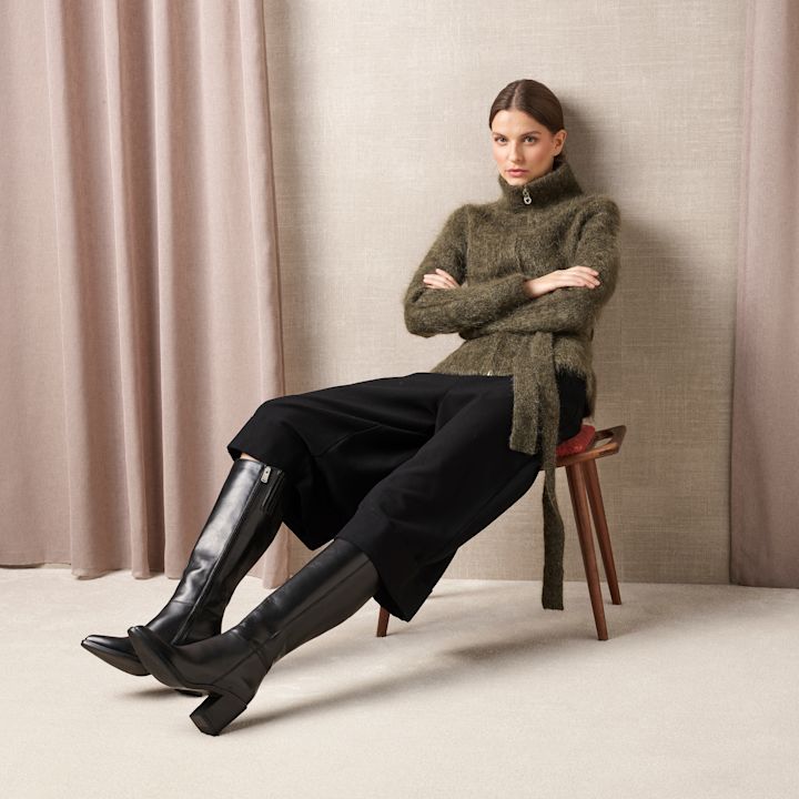 Person i grön stickad tröja och svarta byxor sitter på pall, bär eleganta svarta läderstövlar med klack och sidodragkedja.