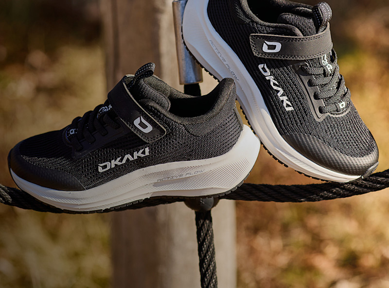 Svarta OKAKI sportskor med kardborreband och vit sula, placerade på ett rep utomhus i naturlig miljö.