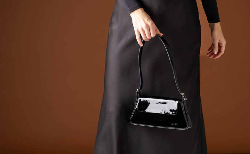 Person i svart klänning håller en liten svart lackhandväska med kort handtag mot brun bakgrund.