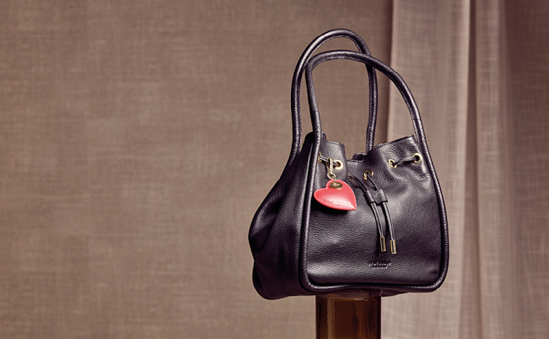 Elegant svart skinnveske med lange håndtak og en rød hjerteformet nøkkelring, plassert mot en nøytral beige bakgrunn.