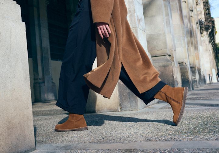 Person i kamelfarget kåpe og mørke bukser går langs steinbygning iført brune semsket ankelstøvletter med flat såle.