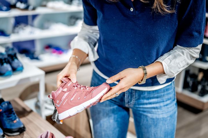 Person i blå tröja håller rosa sportskor med vit sula i en skobutik. Flera andra skor syns på hyllor i bakgrunden.
