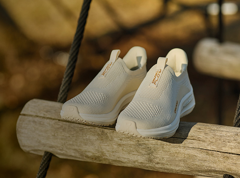 Beige sneakers från Okaki med stickad ovandel och vit sula, placerade på en träbalk utomhus.