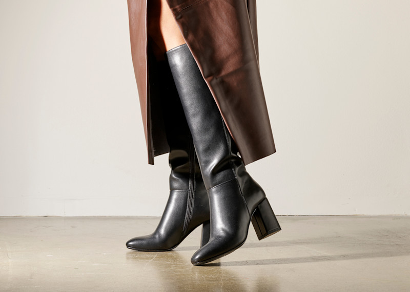 Elegante svarte støvletter med blokkert hæl under en brun skinnkjole, vist mot en nøytral bakgrunn.