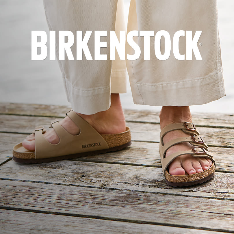 Beige Birkenstock sandaler med korksula på trätrall, burna med ljusa byxor. Varumärkesnamnet syns i vitt överst.