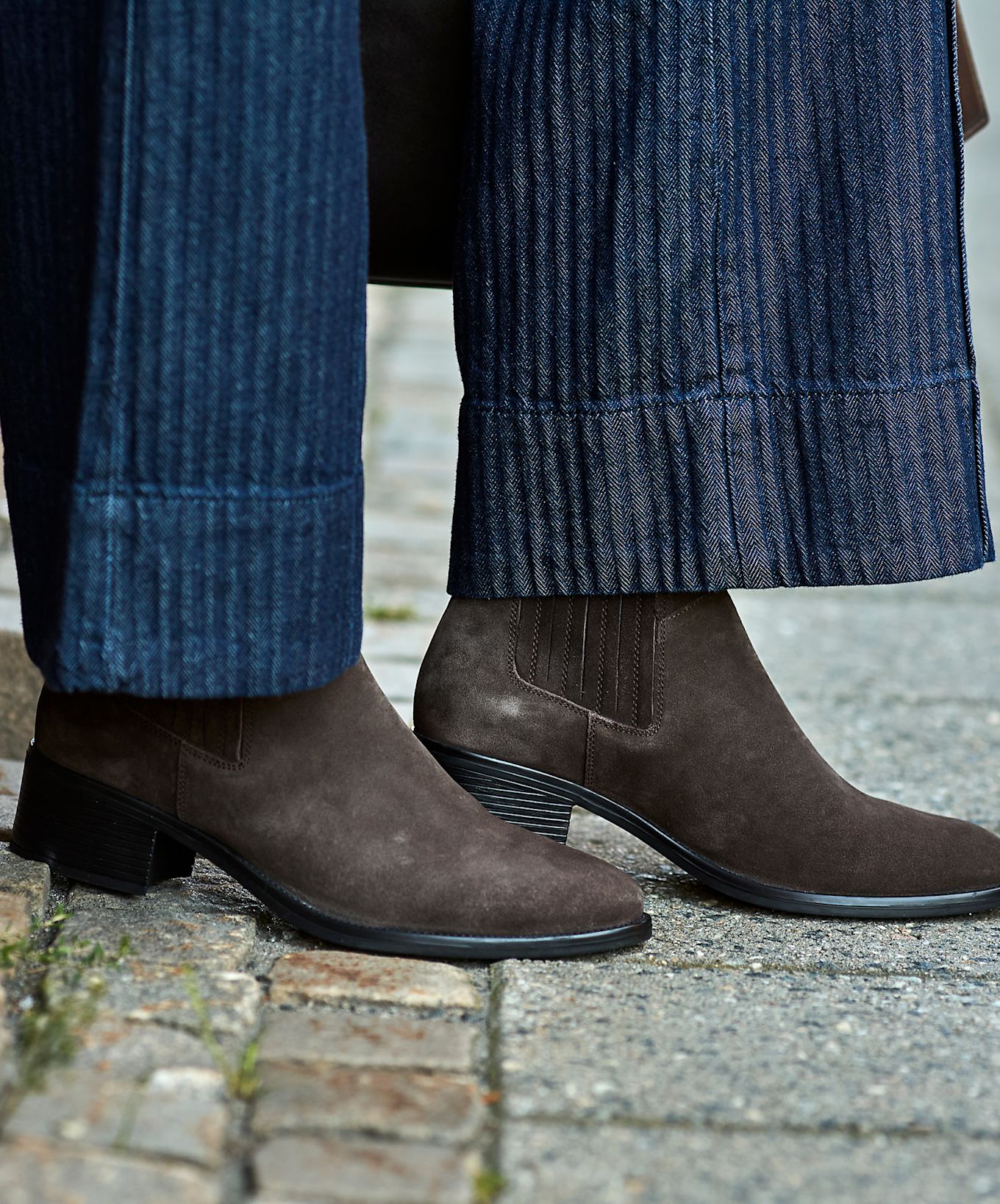 Bruna mockaboots med elastiska sidor under mörkblå randiga byxor, stilrena chelsea-stövlar med låg klack på stenbelagd mark.