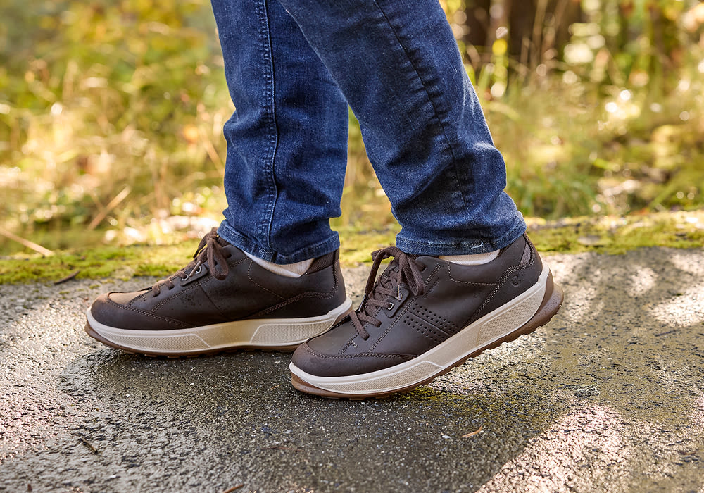 Brune sportslige lærsneakers med beige såle, brukt med mørke jeans på en sti i naturen.