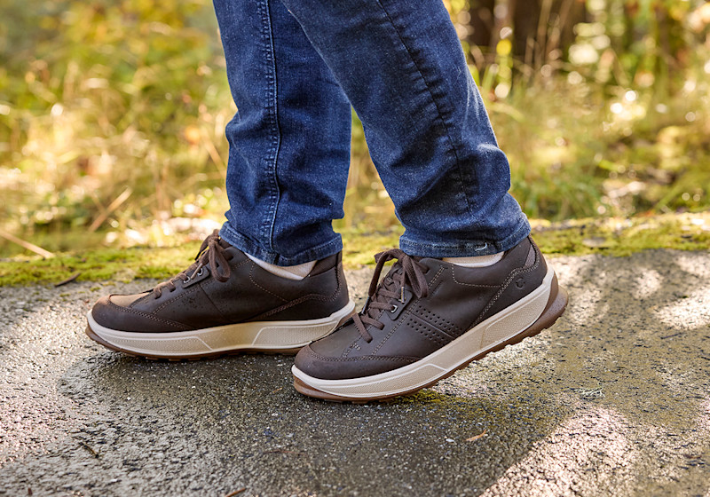 Brune sportslige lærsneakers med beige såle, brukt med mørke jeans på en sti i naturen.