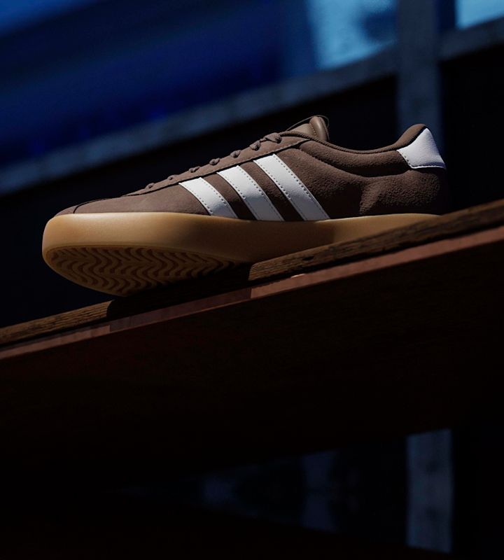 Brun adidas sneaker i mockasstil med vita three stripes och beige gummisula, placerad på en träyta i mörk miljö.