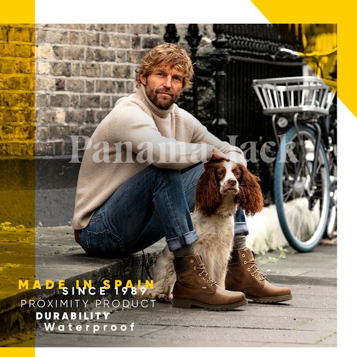 Man i beige tröja och jeans sitter på trottoaren vid en tegelvägg och klappar en brun och vit hund, med bruna vattentäta stövlar, cykel i bakgrunden.