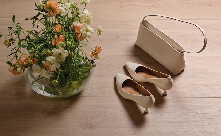 Beige eleganta pumps med spetsig tå och klack, matchande handväska och blombukett på trägolv.