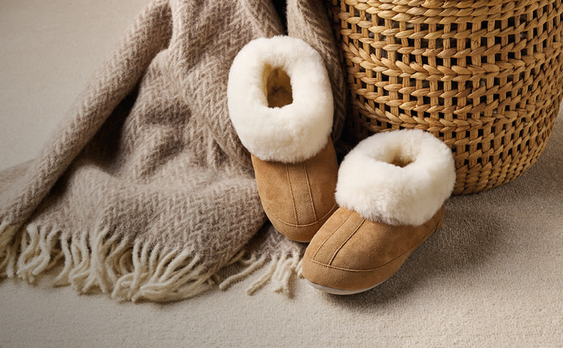 Ljusbruna tofflor med vit fluffig pälskant bredvid en beige filt och flätad korg på en ljus matta.