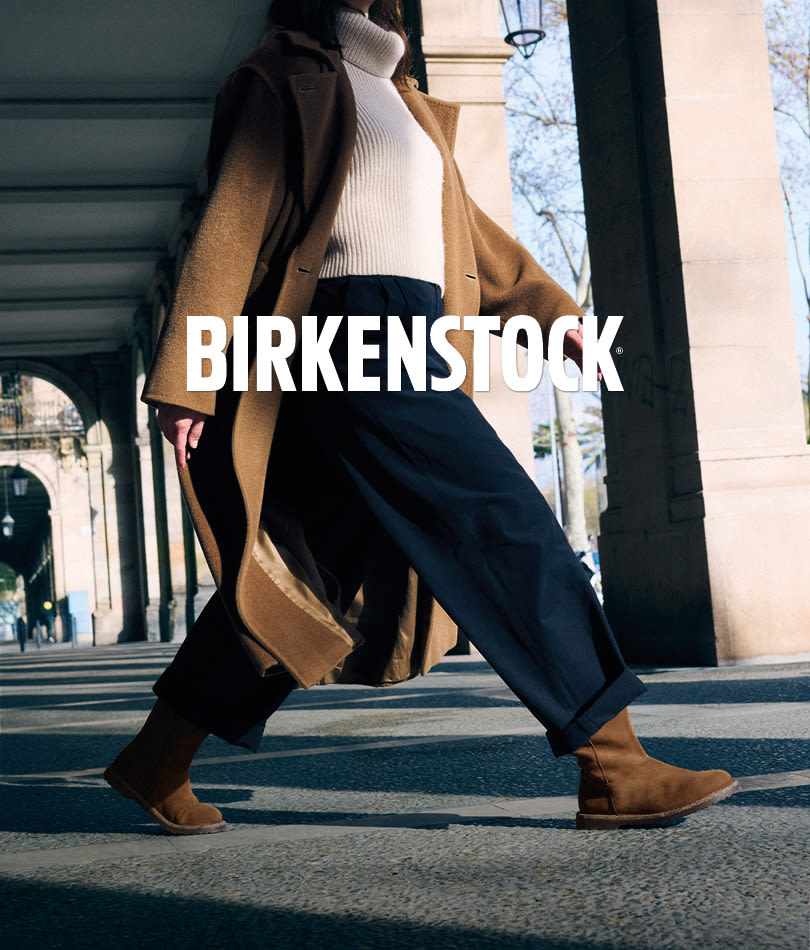 Person i kamelfarget kåpe og mørke bukser går med brune Birkenstock ankelstøvletter i semsket skinn under en arkade.
