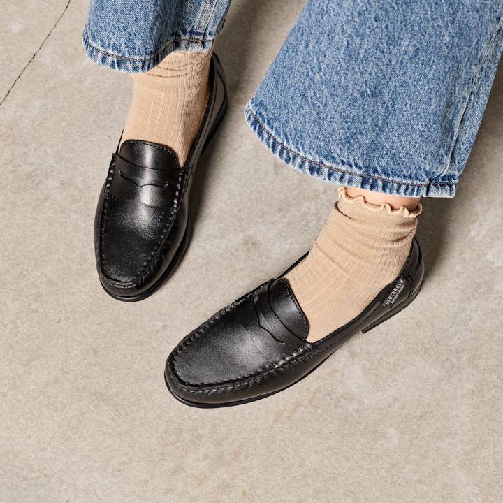 Svarta klassiska loafers i läder med beiga strumpor och blå jeans, fotograferade på betonggolv.