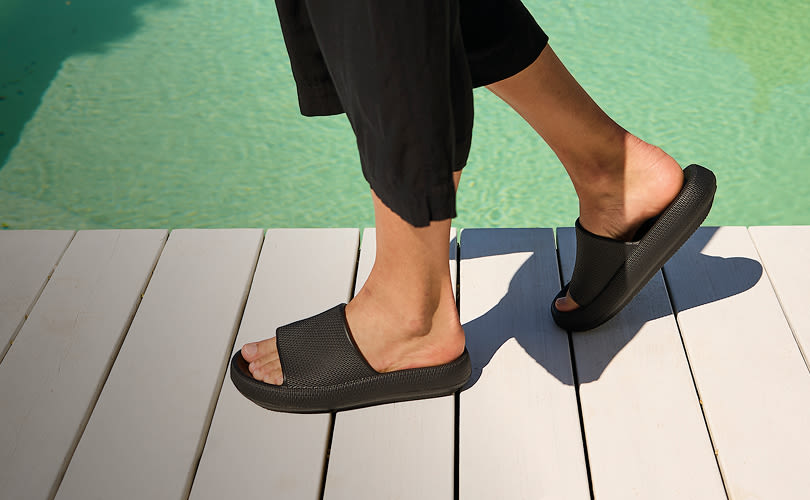 Person med svarte shorts går i svarte, slippers fra Unified på hvit tredekk ved turkis basseng.