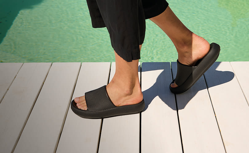 Person med svarte shorts går i svarte, slippers fra Unified på hvit tredekk ved turkis basseng.