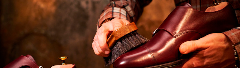 Händer putsar en brun lädersko med en borste i ett varmt upplyst rum.