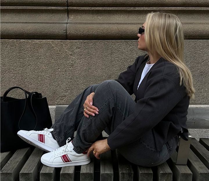 Kvinna med blont hår och solglasögon sitter på en bänk vid en stenmur, iklädd svart jacka, grå byxor och vita sneakers med röda detaljer, med en svart väska bredvid sig.