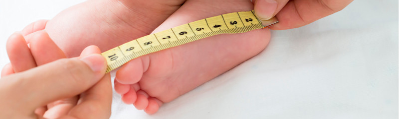 En person måler lengden på en babys fot med et gult målebånd mens babyen ligger på en hvit overflate.