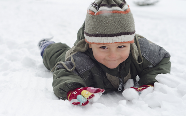 Barn som ligger på magen i snön iklädd grön vinterjacka, randig mössa och färgglada vantar, leende och leker i snön.