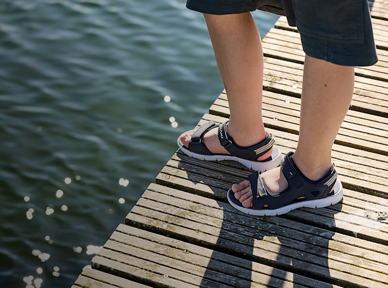 Barn står på en brygge ved vann iført marineblå sportsandaler med hvit sål og justerbare stropper.