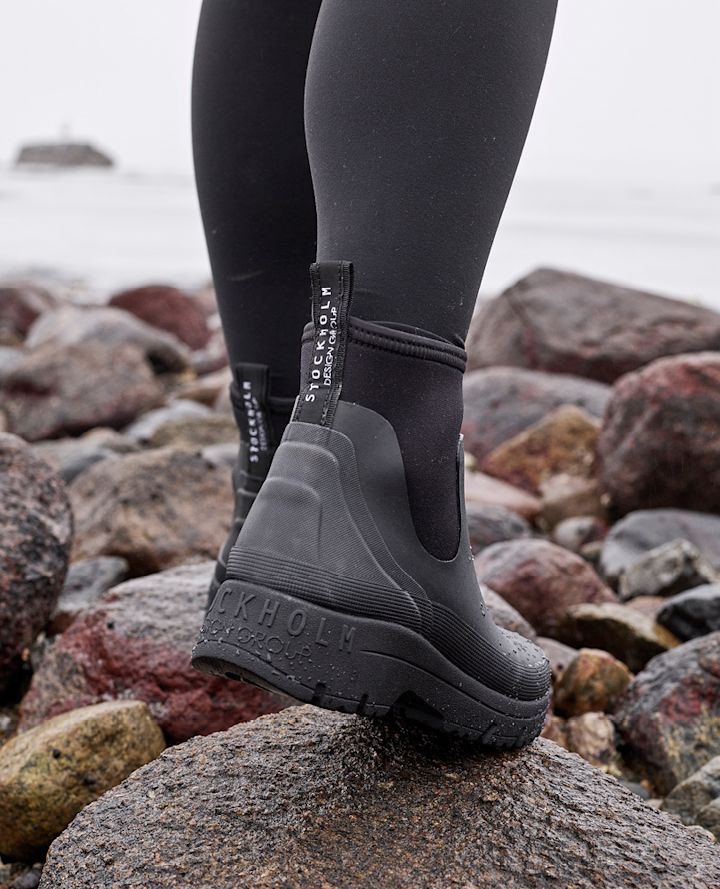 Person med svarte Stockholm Design Group-støvler står på steinete strand nær sjøen i overskyet vær.