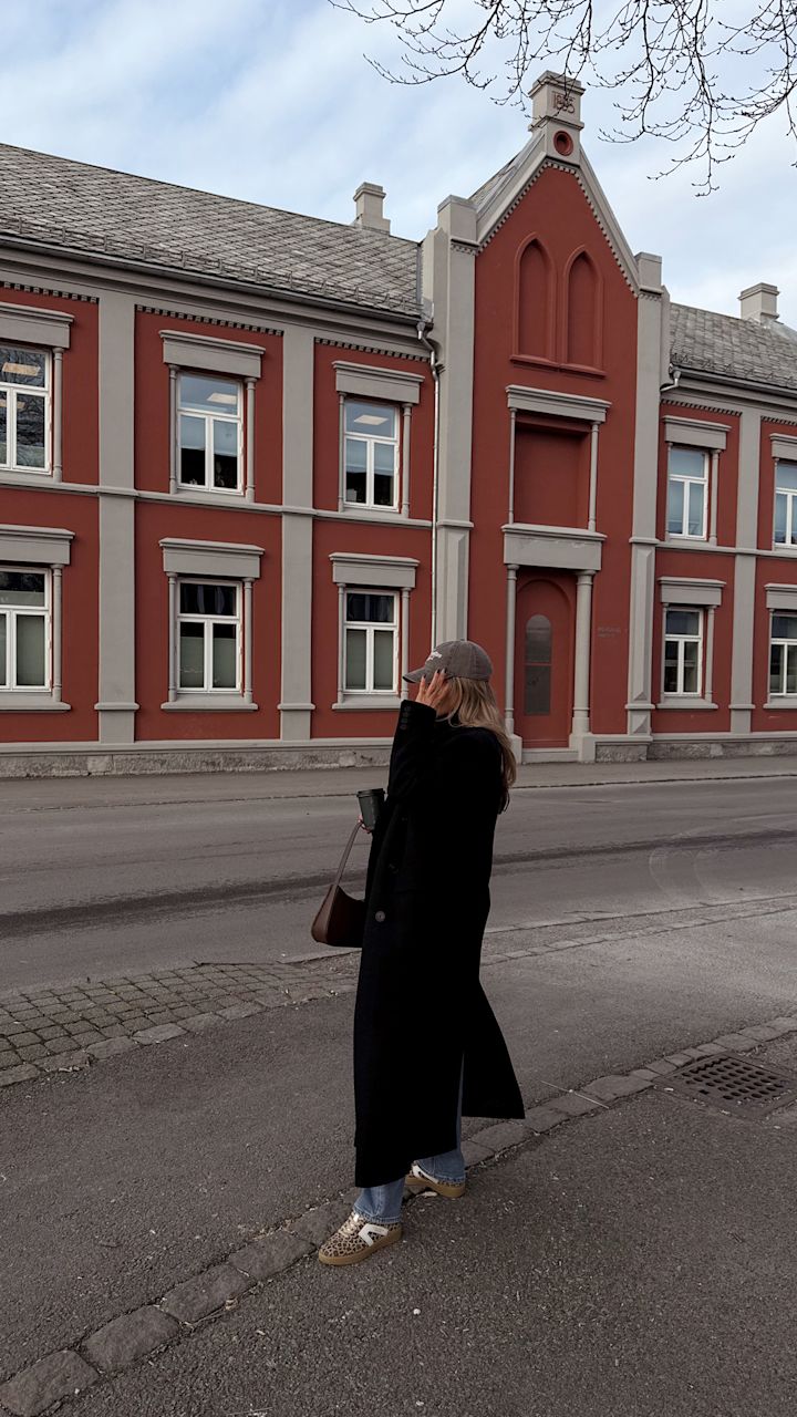 Kvinna i lång svart kappa, blå jeans, leopardmönstrade skor och grå keps håller en kaffekopp och står på trottoaren framför en röd byggnad med vita fönsterkarmar.