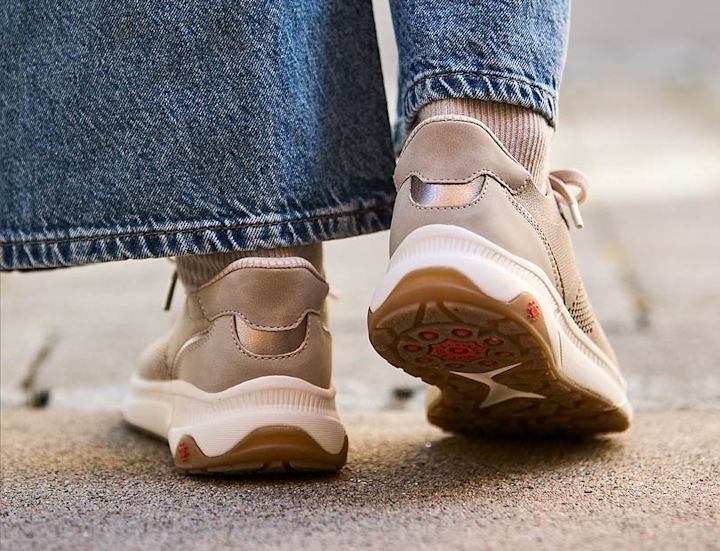 Beige/rosa sneakers med hvit såle og røde detaljer, sett bakfra på en person i jeans som går utendørs.