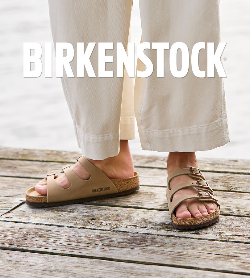 Beige Birkenstock sandaler med korksåle og spenner, brukt med lyse bukser på tredekk ved vann. BIRKENSTOCK-logo vises.
