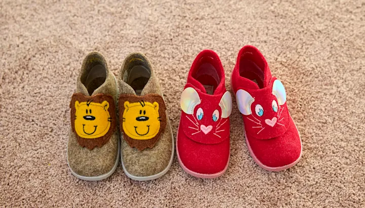 To par barnetøfler: beige med løvefigurer og røde med musemotiv, plassert på beige teppe.