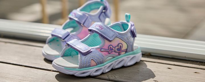 Barnsandaler i lila och rosa med hjärt- och nyckeldetaljer placerade på ett ljust trägolv i solljus.