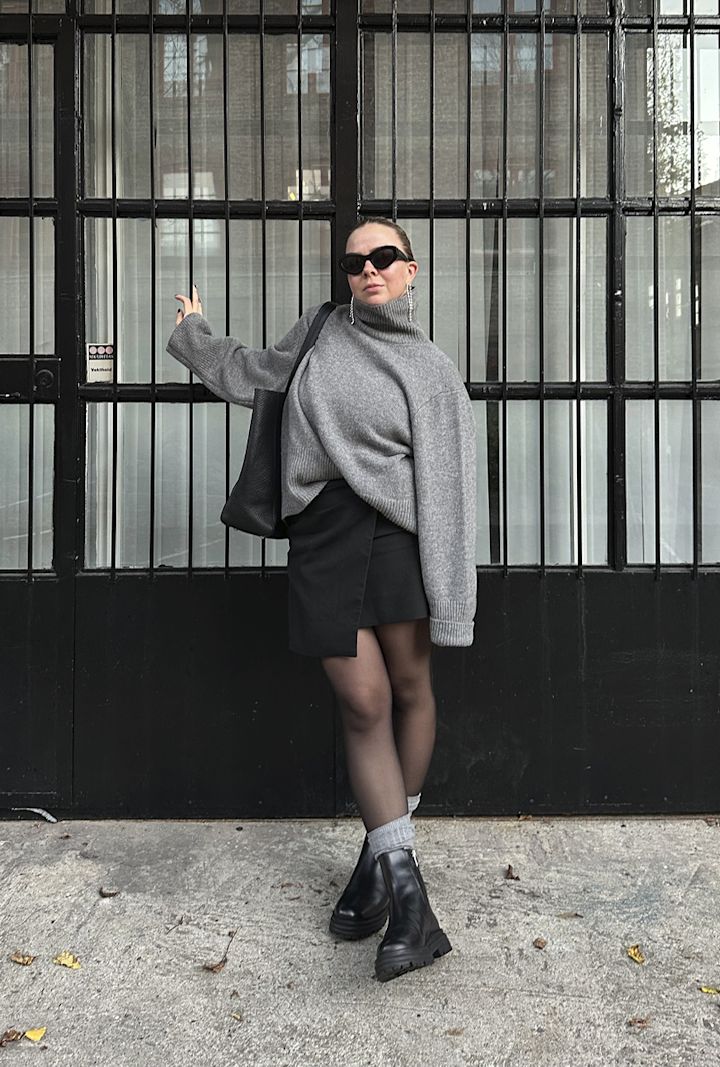 Kvinna bär en grå polotröja, svart kjol, svarta stövlar och solglasögon, står framför en svart gallergrind med glasrutor och håller en svart väska över axeln.