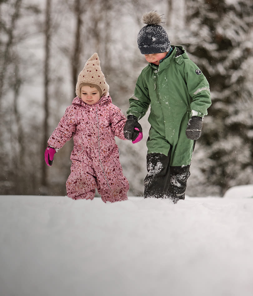 To barn i vinterdresser, en i rosa og en i grønn dress, går hånd i hånd gjennom dyp snø i en vinterskog.