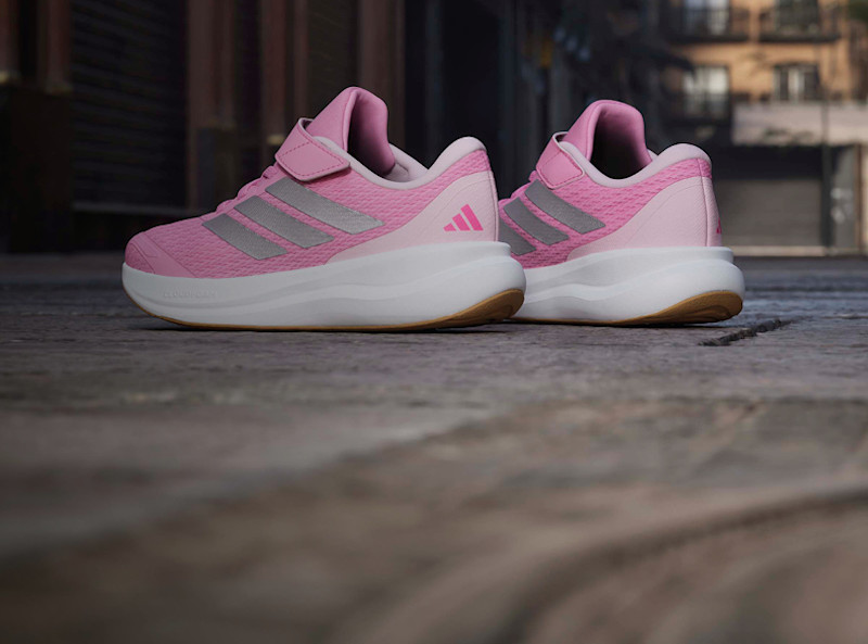 Rosa Adidas sneakers med kardborreband, grå tre-ränder, vit sula och gummibotten, sedda bakifrån på ett stengolv.