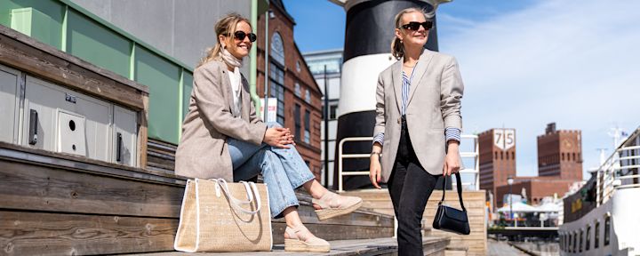 To personer i beige blazere i bymiljø, en sittende med beige espadrillos og en stående med solbriller og veske.