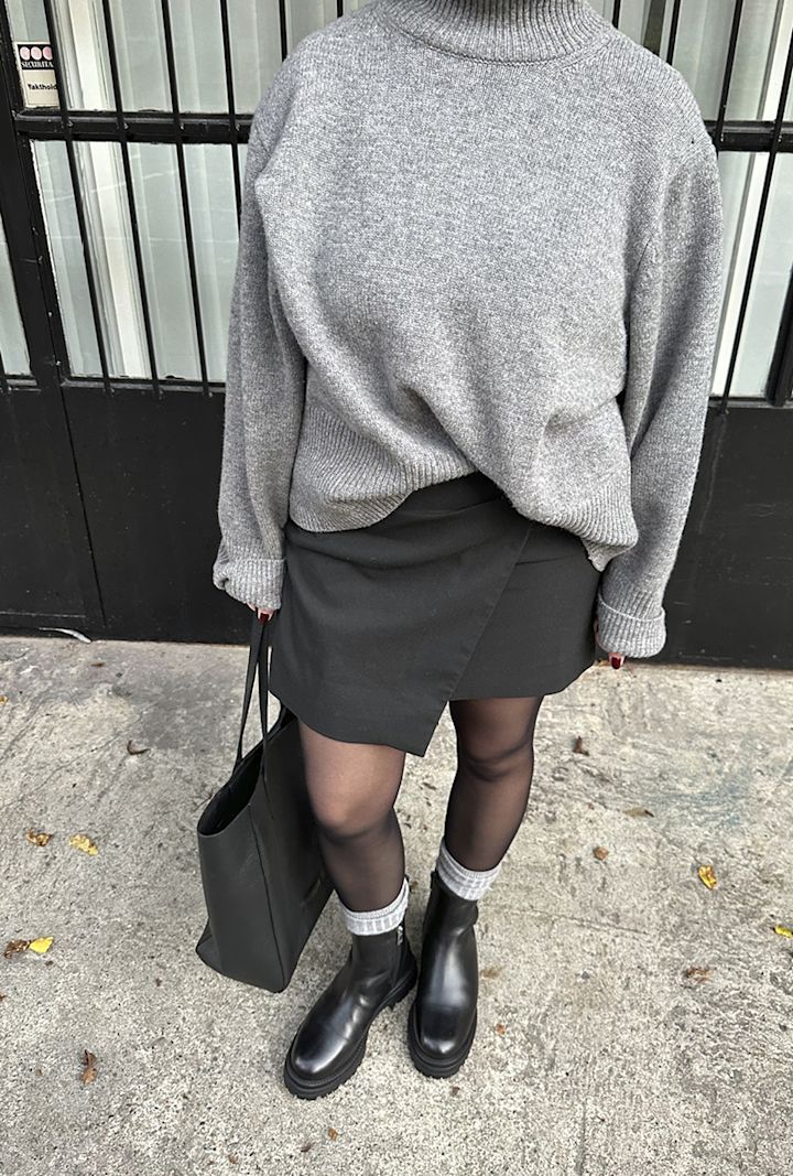 Person klädd i grå tröja, svart kjol, svarta stövlar med gråa strumpor och håller en svart väska, står på trottoaren framför en svart dörr med glasrutor.