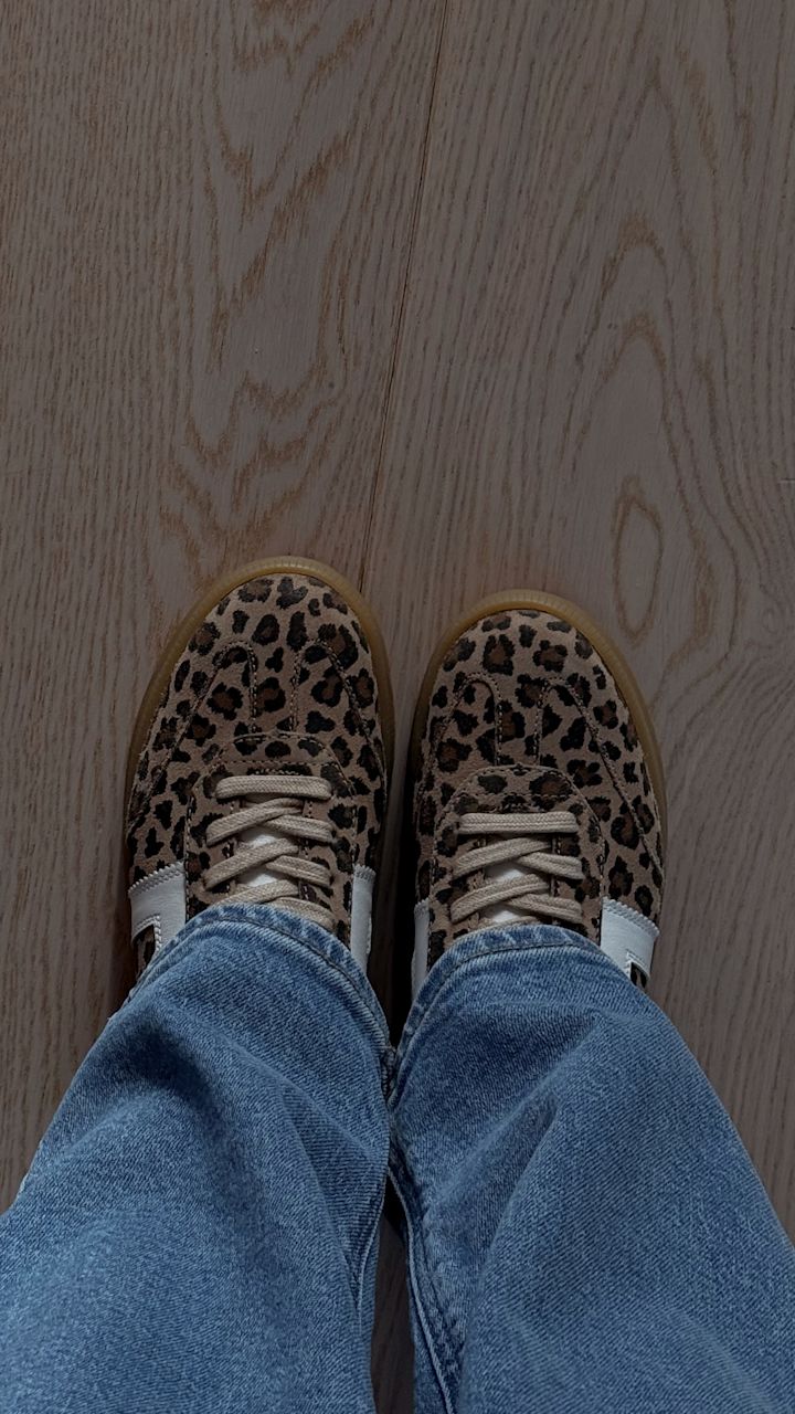 Skor med leopardmönster och vita detaljer på fötterna på en person som bär blå jeans, stående på ett ljust trägolv.