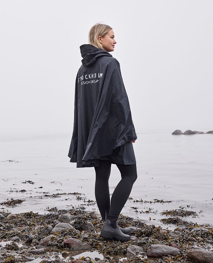 Kvinne iført mørk regnponcho med teksten "STOCKHOLM DESIGN GROUP" står på steinete strand ved sjøen i tåke.