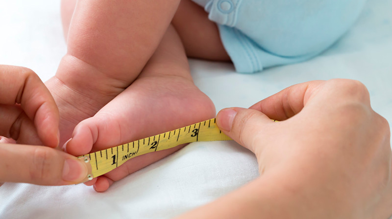 Händer mäter en babys fot med ett gult måttband på en vit yta.