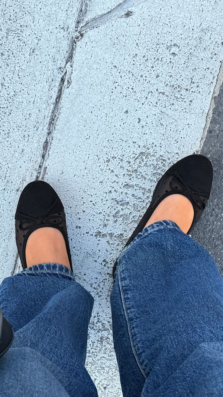 Sorte ballerina sko med små prikker og sløyfedetalj på føttene til en person iført blå jeans, stående på en lys grå betongoverflate.