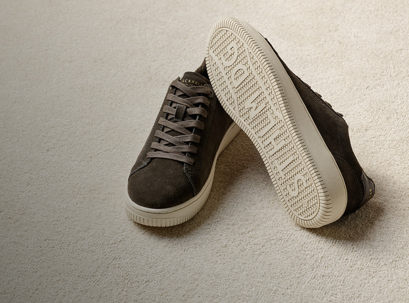 Bruna mocka sneakers med snörning och vit gummisula från Stockholm Design Group, placerade på en ljus matta med en sko vänd för att visa sulan.