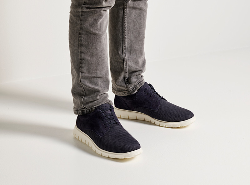 Person med grå jeans står på vitt golv och har på sig mörkblå sneakers med vita sulor.