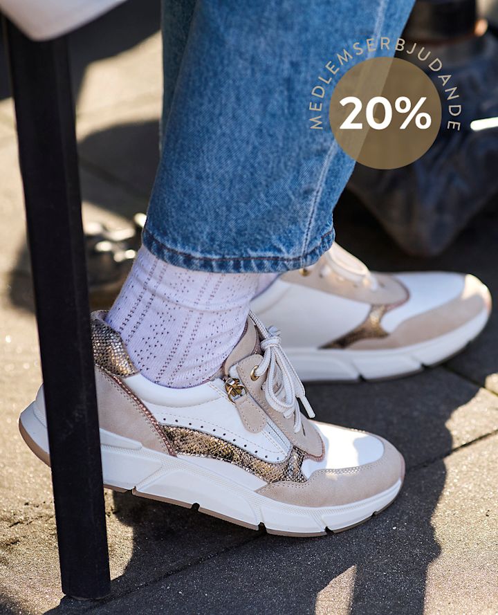 Person med vita och beige sneakers med ormmönster sitter utomhus, iförd vita strumpor och blå jeans. Cirkel med texten "MEDLEMSTILLBUD 20%".