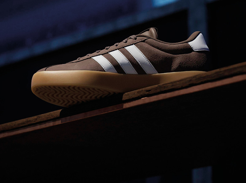 Brun Adidas sneaker i mocka med vita ränder och gummisåla, placerad på en trälist mot mörk bakgrund.