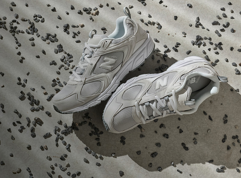 Hvite New Balance løpesko med mesh-detaljer plassert på grå betongoverflate med små steinbiter spredt rundt.