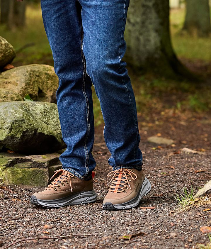 Person i blå jeans bär ljusbruna vandringsskor från Okaki med tjock grå sula på en skogsstig omgiven av stenar.