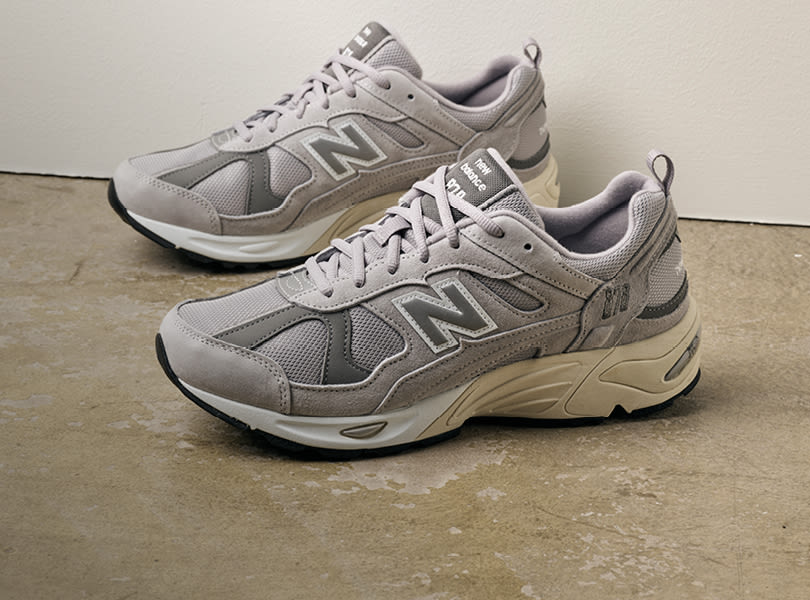 Grå New Balance løpesko med mesh og semsket skinn, tykk såle og NB-logo på siden, plassert på betonggulv.