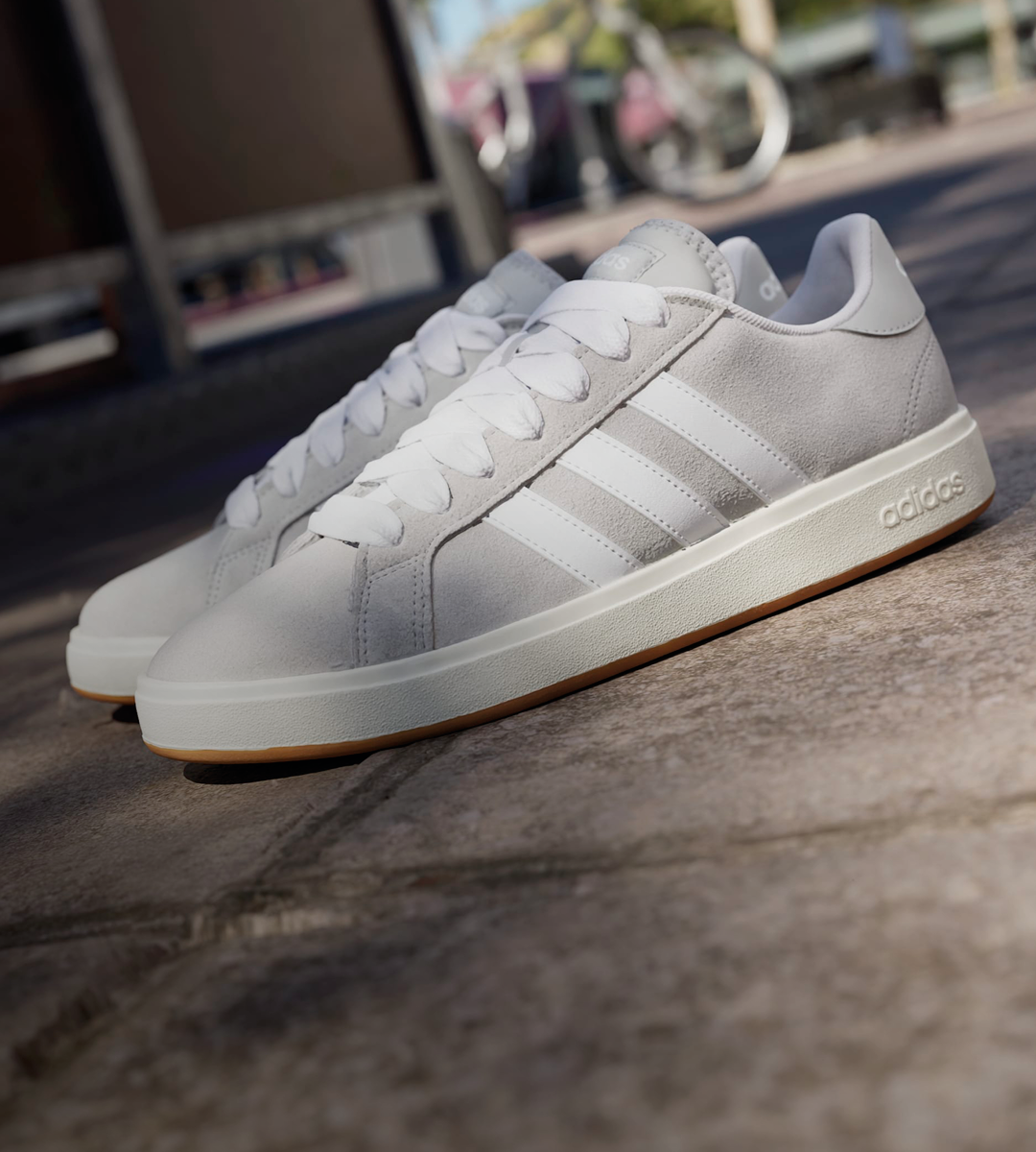 Hvite Adidas sneakers med semsket overflate, tre striper på siden og gummiaktig såle, plassert på en betongoverflate utendørs.